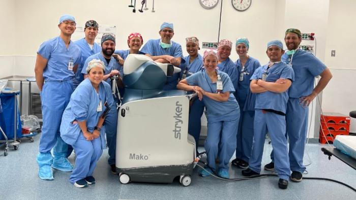 Twelve doctors in nurses in blue scrubs stand in a brightly lit operating room standing around a robot used for operating.