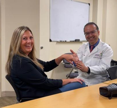 A man in a white lab coat holds up a woman's left hand to show off her wedding ring, they are both sitting in a conference room at a long, brown table. 