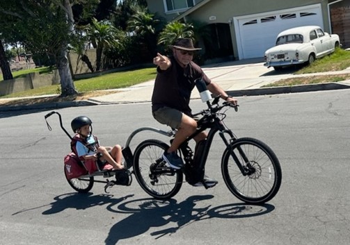 Middle-aged man in a hiking, wide brim hat, khaki shorts and Hoka shoes rides a bike down the street with his son riding on an attachable seat behind him 