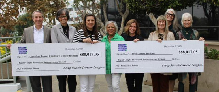 Eight people holding two giant checks while standing in front of some trees to the side of a small staircase. 