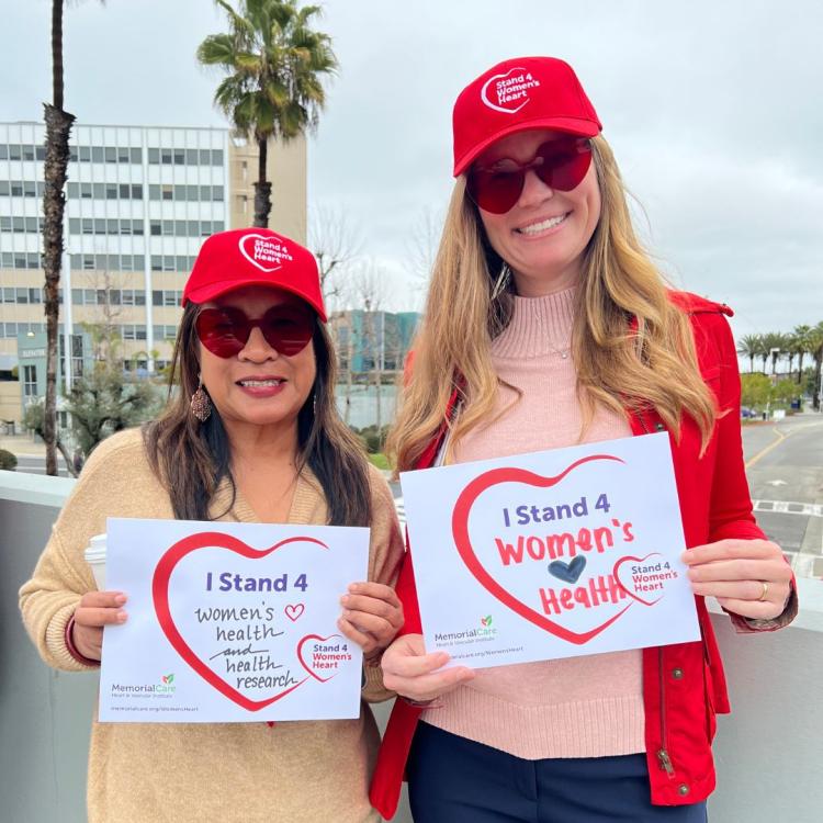 Two women wearing red hats and red heart sunglasses holding signs that say "I Stand 4 Women's Heart Health"