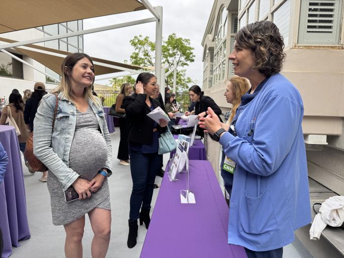 A pregnant woman in a grey dress and grey denim jacket talks to a nurse in blue scrubs over a table with a purple tablecloth. 