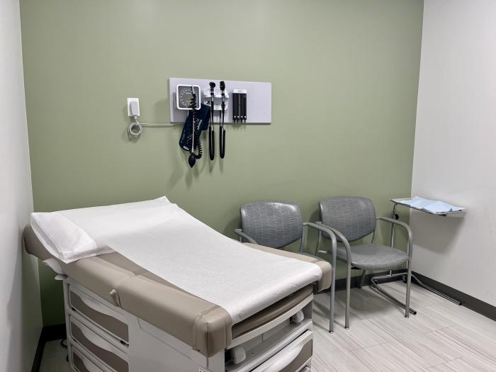 A medical exam room with a teal and white wall furnished with a patient bed, two grey chairs, a blood pressure monitor, and eye and throat instruments. 