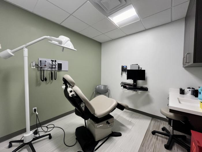 A medical exam room with a teal and white wall furnished with a patient chair, overhead light, blood pressure monitor, and computer.