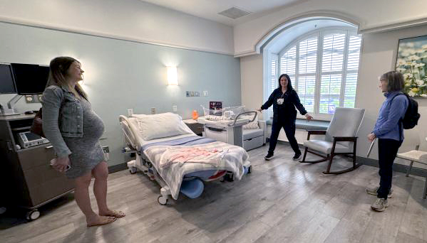 A pregnant woman in a grey dress and grey denim jacket stands in a delivery room with a bed and furniture for family to sit in. 